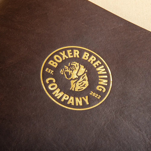 A close up of a gold foil deboss personalisation on the cover of a leather menu cover that has a brushed metallic effect