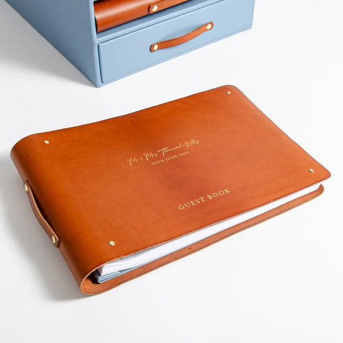 A personalized brown leather wedding guest book binder with gold foil lettering, shown in the foreground with a light blue leather storage case partially visible in the background.