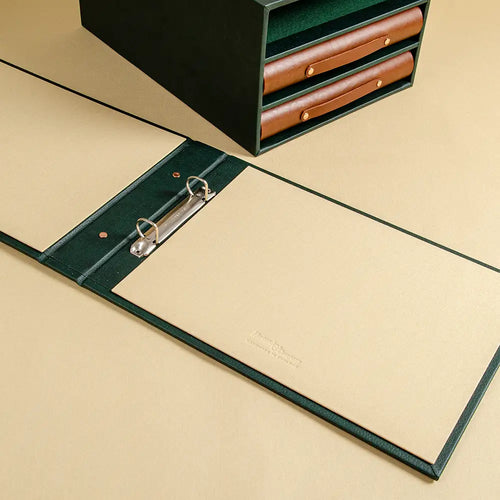 A dark green leather ring binder folder with tan interior and a 2 ring silver metal ring binder mechanism. Behind is a matching organiser slipcase box with brown leather ring binders partially visible in the background.