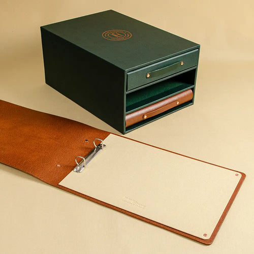 A dark green faux vegan leather slipcase storage box with a brown leather ring binder laying open in front and a green vegan leather and brown leather binder in the slipcase slots