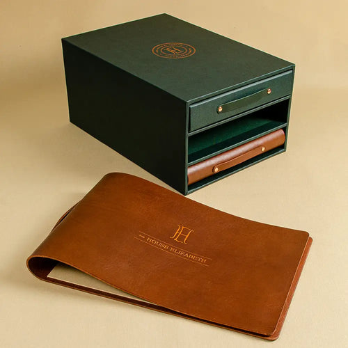 A dark green luxury presentation box with a small front drawer and a brown leather ring binder cover laid in front. Both pieces feature gold monogram and text detailing.