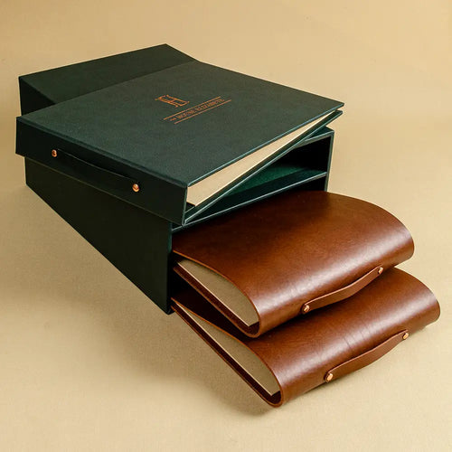 A set of three leather portfolio binders arranged to show both the dark green presentation box and the brown leather covers. The top A4 landscape binder is personalised with a gold foil logo.