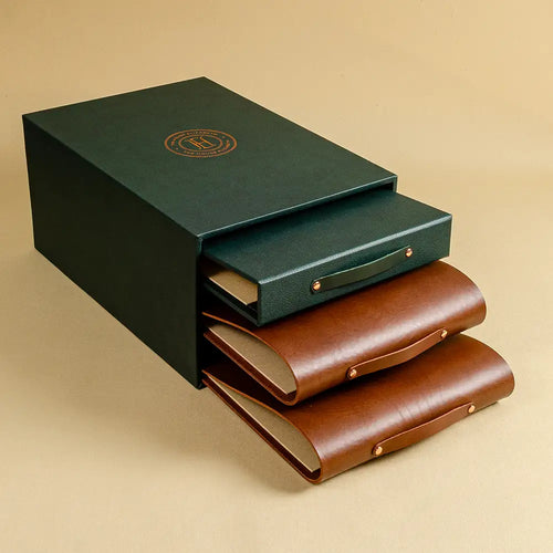 A dark green quality presentation box with a circular gold emblem embossed on top, partially open to reveal two brown leather ring binders nested inside and a green ring binder set against a beige background