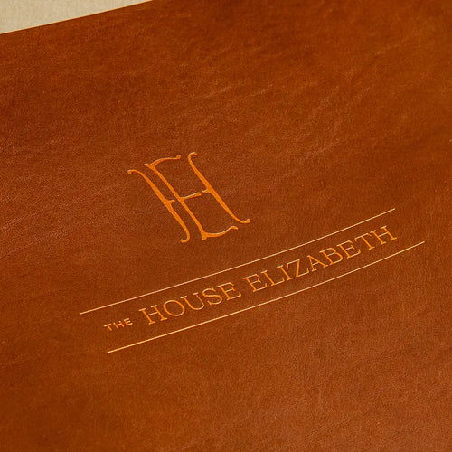 A close-up of a brown textured leather binder with an embossed copper orange monogram and the text 'THE HOUSE ELIZABETH' printed on the cover.