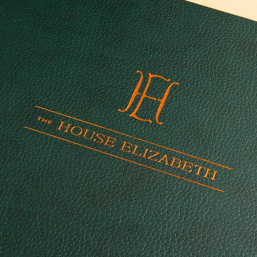Close-up of a dark green faux leather cover with copper gold foil lettering embossed into the cover reading 'THE HOUSE ELIZABETH' and a large ornate 'H' monogram.