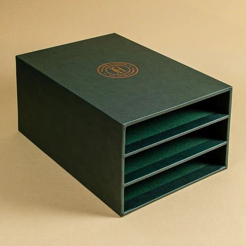 A dark forest green rectangular storage box with three open shelves, featuring a circular gold logo on the top lid, designed to hold 3 books or ring binders.