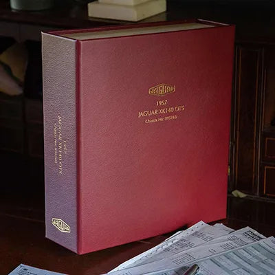 Red leather bound box file with gold foil embossing of a logo and title on the cover and the spine sitting on dark brown desk