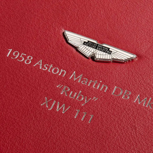 A close-up of a red leather box with embossed silver text reading '1958 Aston Martin DB Mk 