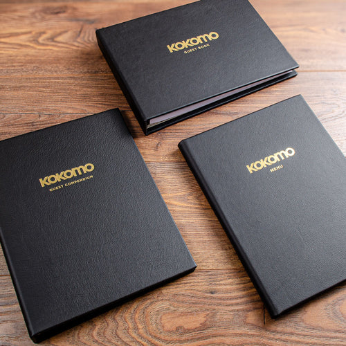 A collection of three black leather binders with the logos of a yacht gold foiled onto the covers and then the titles of; guest compendium, guest book and menu also gold foiled onto the cover 