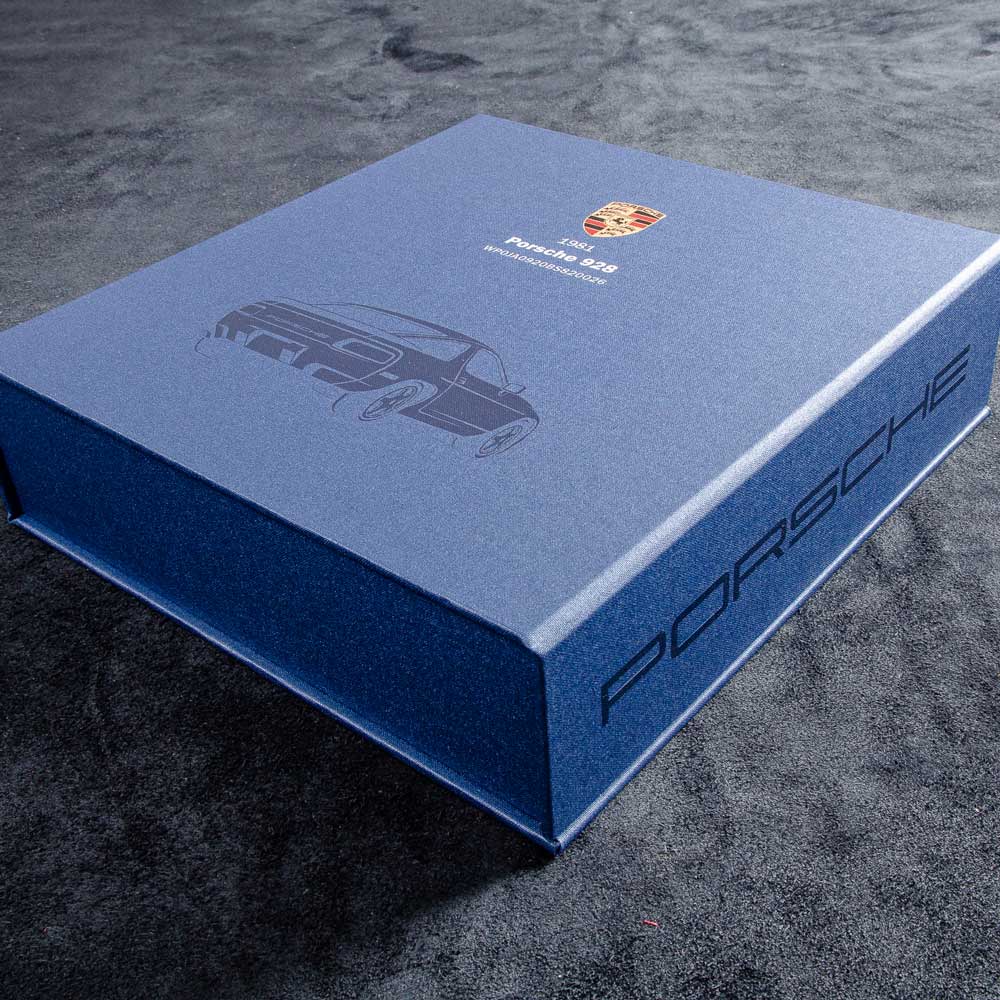 Blue Porsche vehicle history document box file with car 928 illustration on a dark surface - Gallery image 1